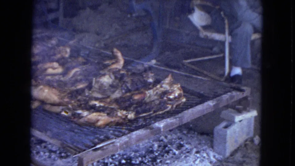 1967 home-movie frame of poultry roasting on a large outdoor grill, with a partially visible adult's legs and feet near the grate.