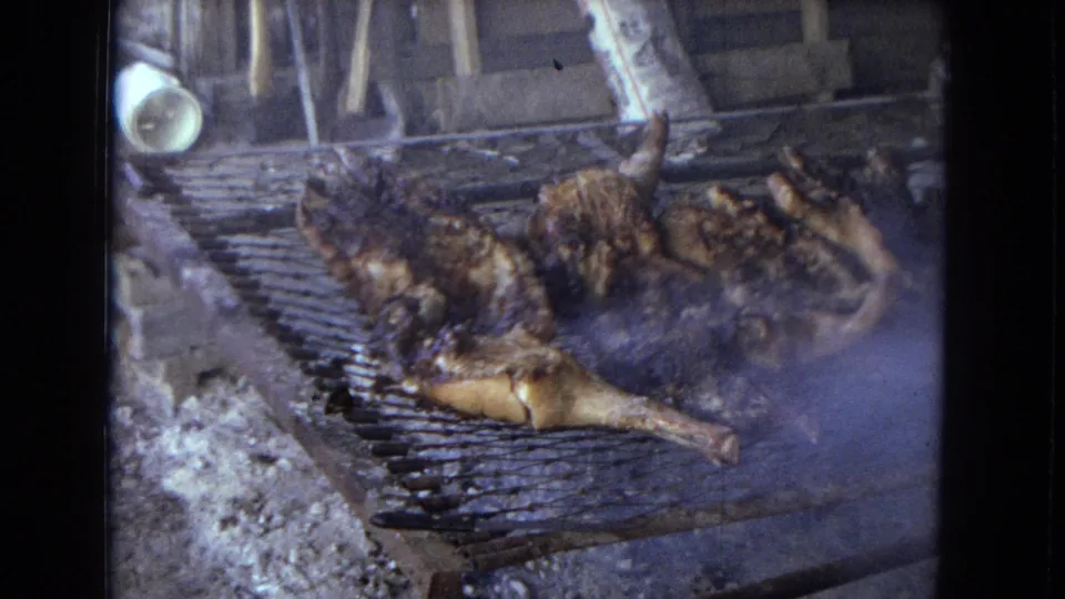 1967 home-movie frame showing browned and charred poultry pieces on a metal outdoor grill, with smoke, ash, and hanging utensils or background objects visible.