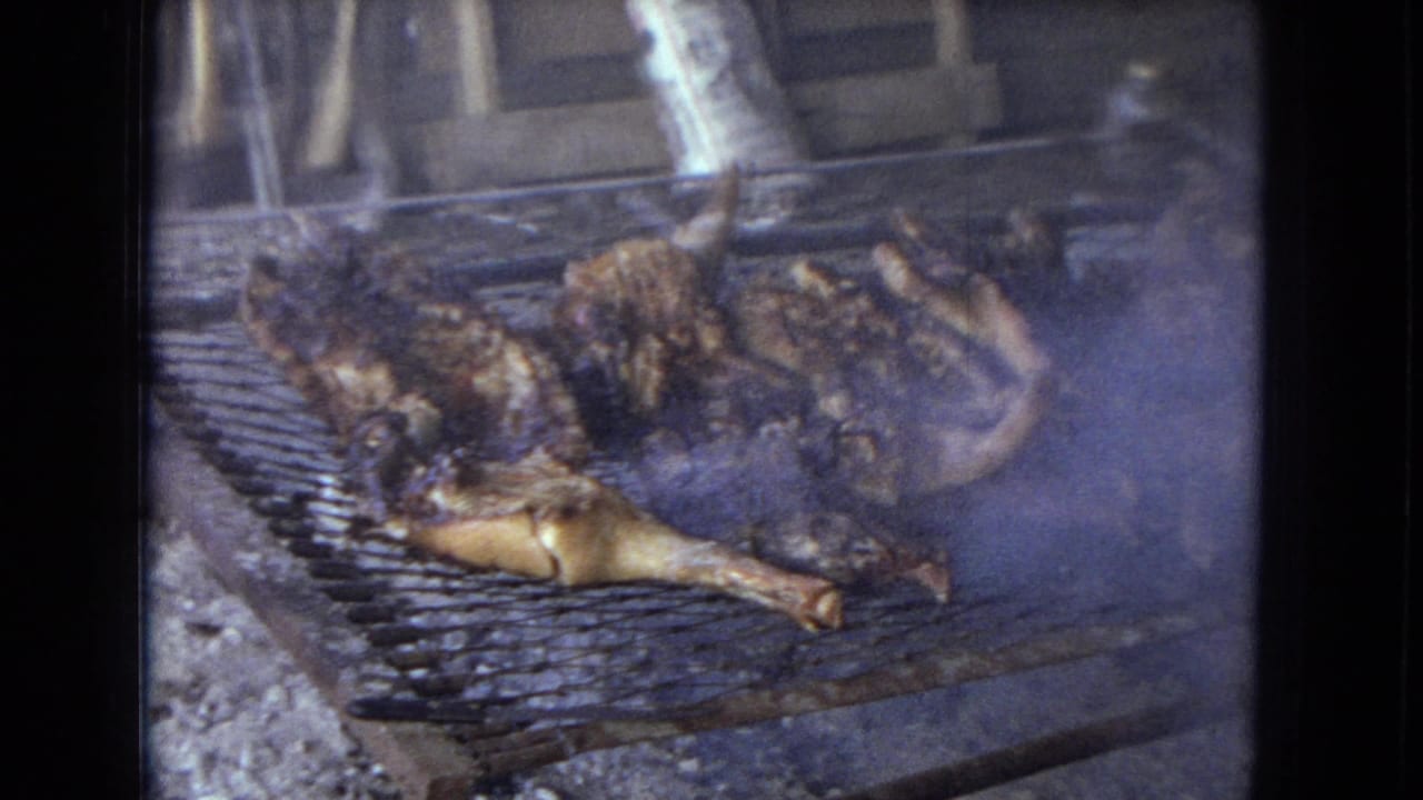 1967 Arizona 8mm frame showing whole chickens roasting over coals on a makeshift wire grill inside or beside a rustic shed.