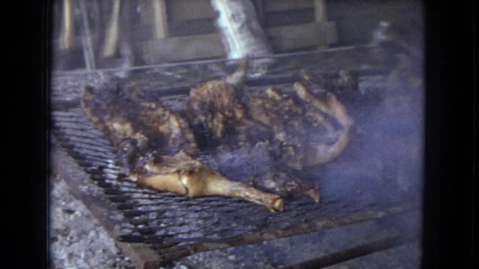 1967 Arizona 8mm frame showing whole chickens roasting over coals on a makeshift wire grill inside or beside a rustic shed.