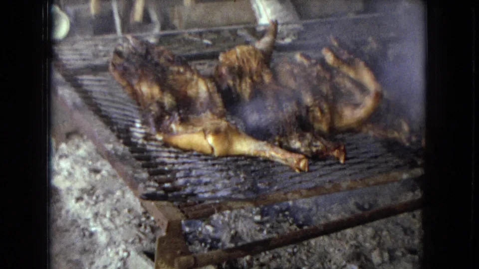 Charred whole chickens sit on a metal grate above ash and coals in a rural outdoor cooking scene.