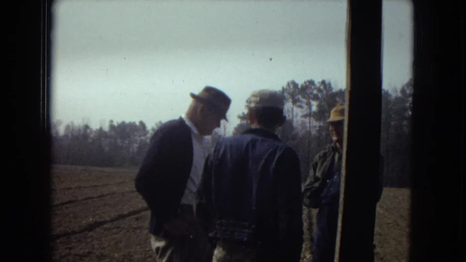 Three adult men, including one in a fedora and suit jacket, stand in conversation near a doorway overlooking a rural field.