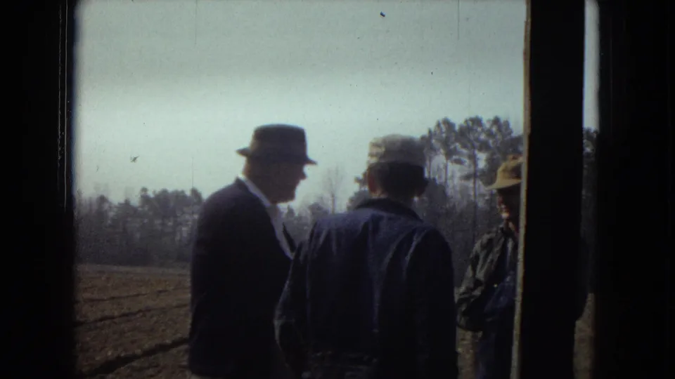 Adult men in hats and jackets gather by a doorway with open farmland and trees visible in the background.