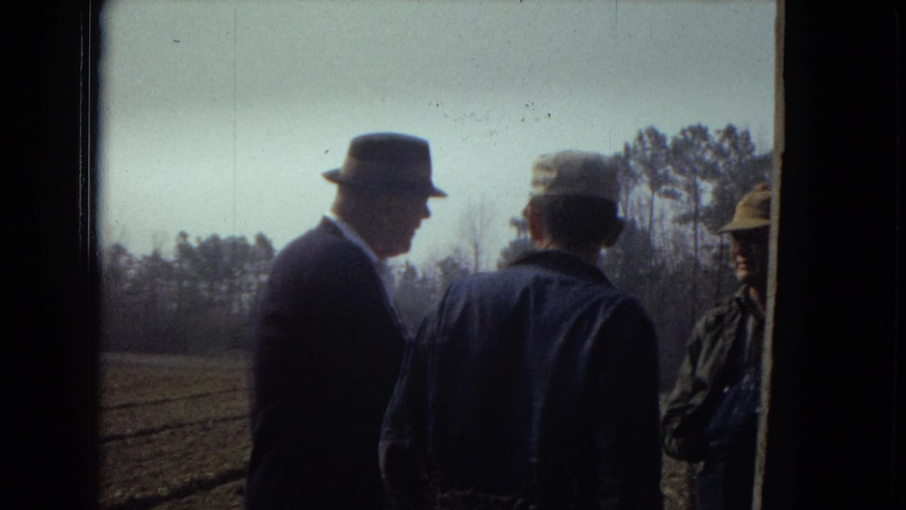 In 1967 home-movie footage, an older man in a dark suit and fedora stands near a plowed rural field with two other adult men, framed by dark edges and a distant tree line.