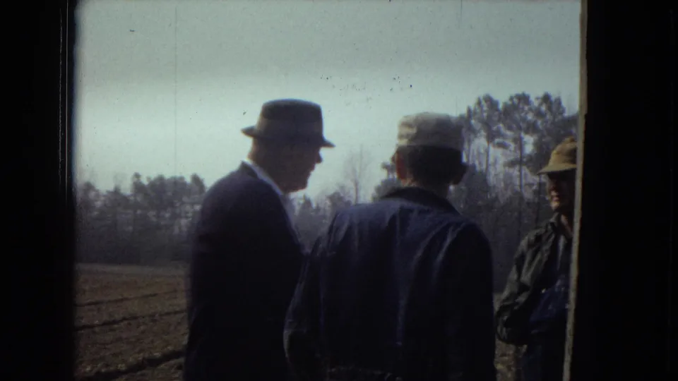 In 1967 home-movie footage, an older man in a dark suit and fedora stands near a plowed rural field with two other adult men, framed by dark edges and a distant tree line.