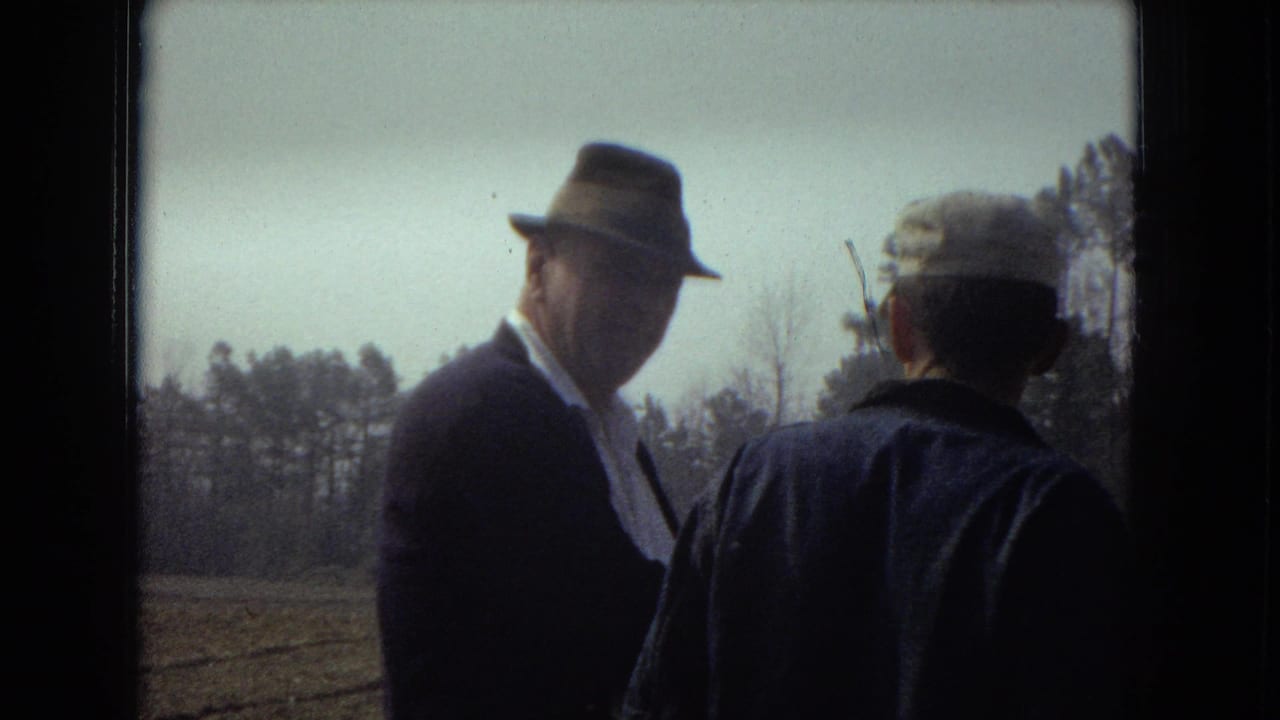 An older man in a fedora turns toward the camera while standing beside a man in a cap near a field.