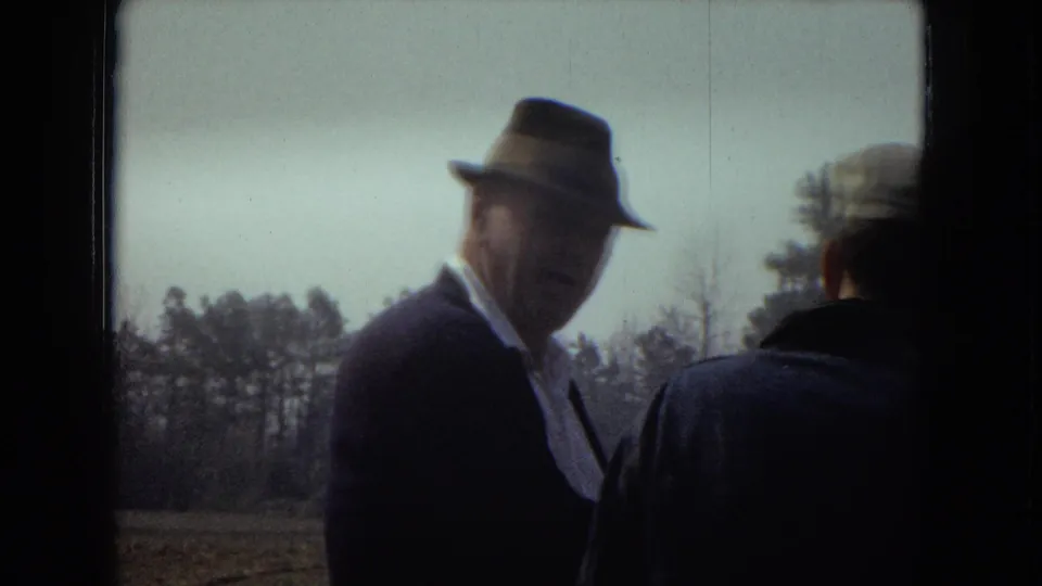 An older adult man in a fedora and dark suit is seen closer to the camera while another man in a cap faces away, with trees and pale sky behind them.