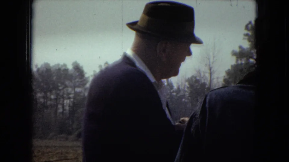 The older suited man appears in profile beside another adult man at the field edge, with the background tree line and dark film borders visible in the frame.