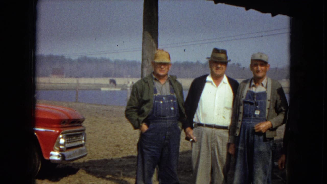 In 1967 United States home-movie footage, three men in workwear and hats stand near a lake or reservoir while the front of a red vintage truck appears at the left edge.