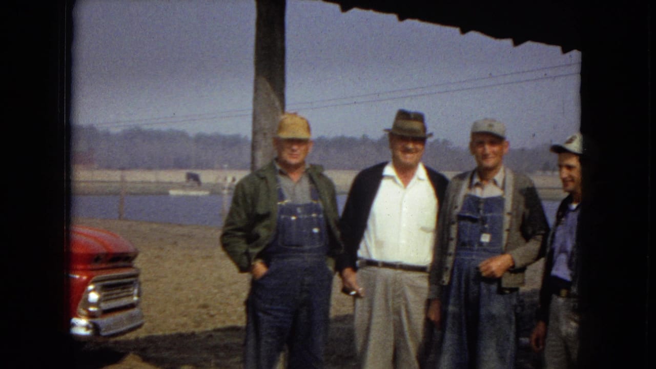 Four men stand in a row outdoors with a lake and red truck partly visible.
