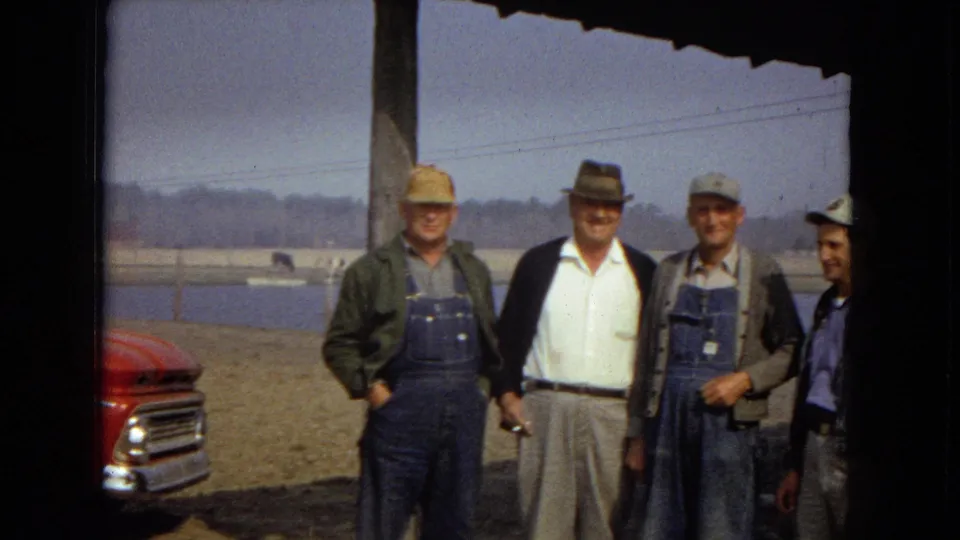 Four men stand together for a casual photograph near a lake or reservoir, with workwear, caps, a fedora-style hat, and part of a red truck visible in the 1967 home-movie frame.