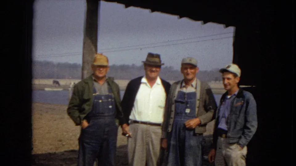 Four men in practical 1960s clothing pose under the edge of a shaded structure near a lake or reservoir, with the red truck mostly out of frame at left.
