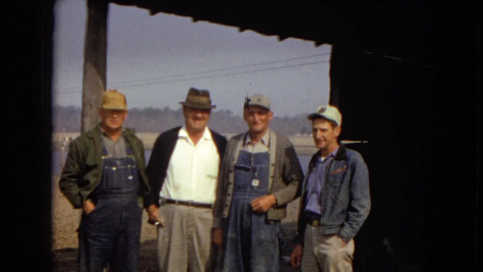 The final frame shows four men facing the camera near a lake or reservoir, with denim workwear, hats, a distant tree line, and overhead shade visible in the 1967 home-movie scene.