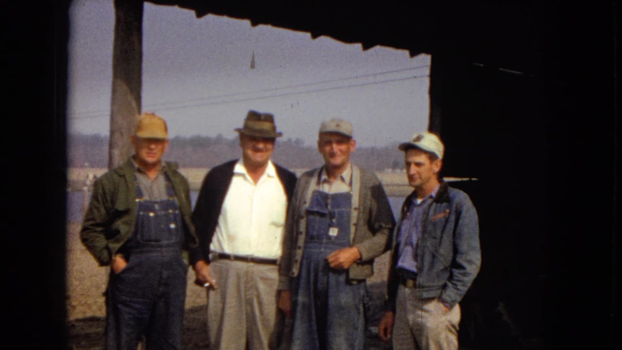 Four older men in work clothes and hats stand together under a roof overhang in 1967 Arizona home-movie footage, with water, power lines, and dirt ground behind them.