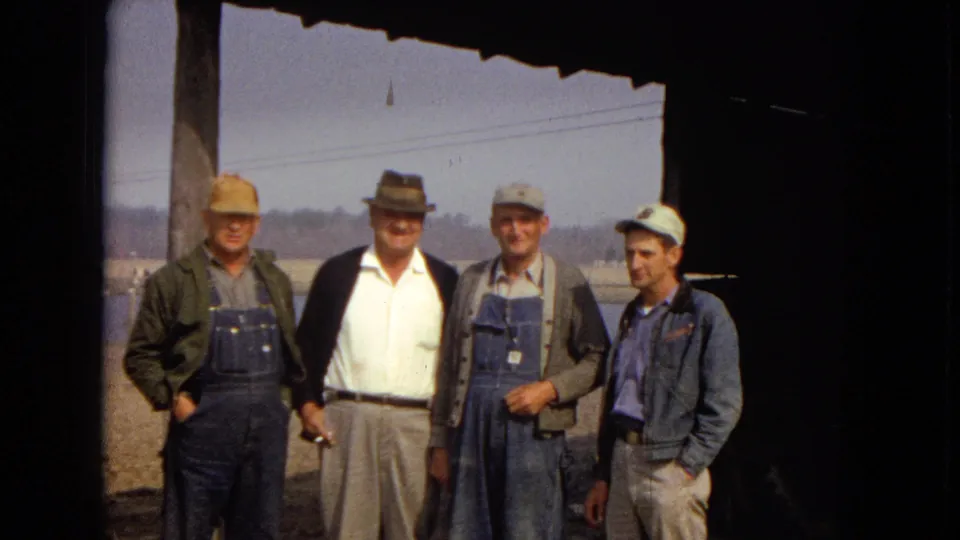 Four older men in work clothes and hats stand together under a roof overhang in 1967 Arizona home-movie footage, with water, power lines, and dirt ground behind them.
