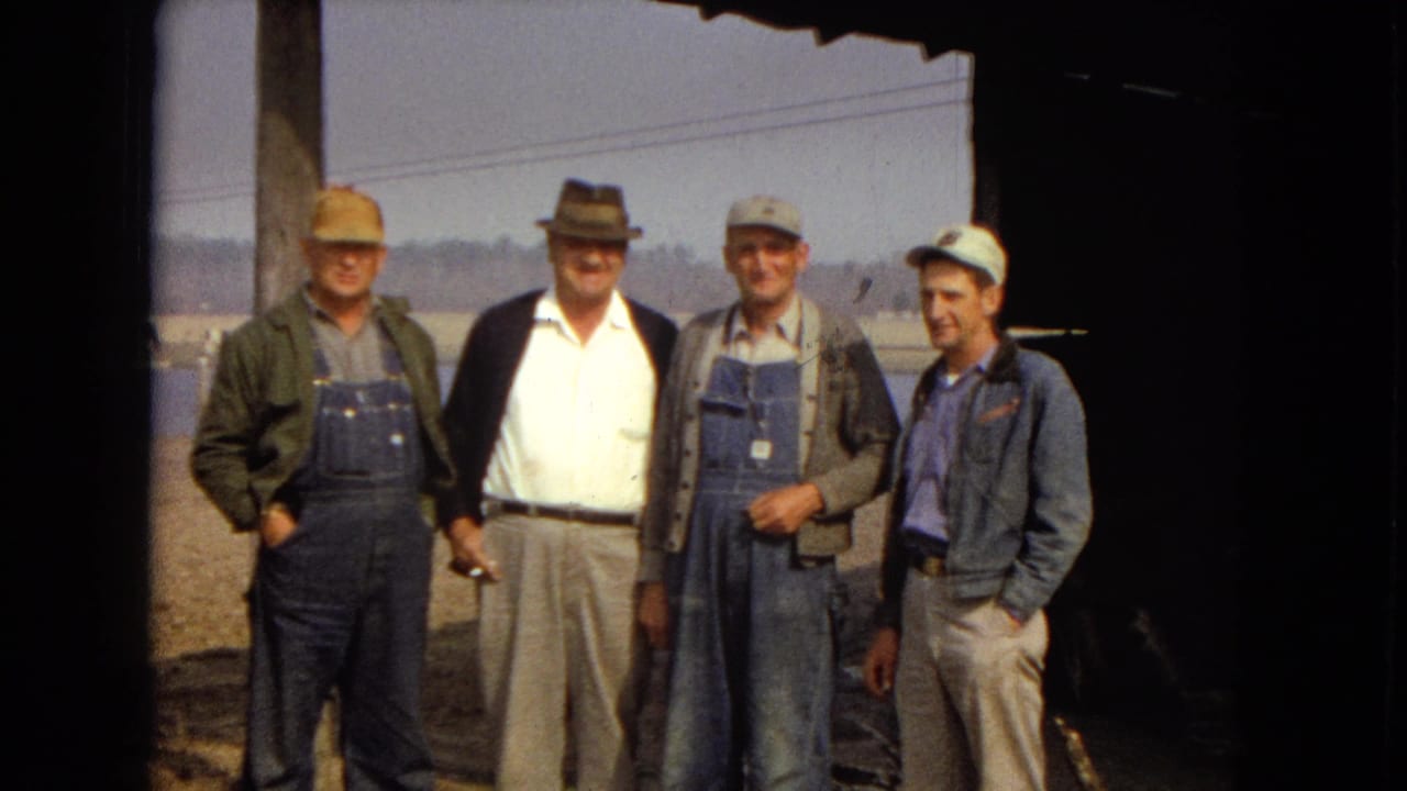 Four men in hats and work clothes stand together beneath a shaded roof edge.