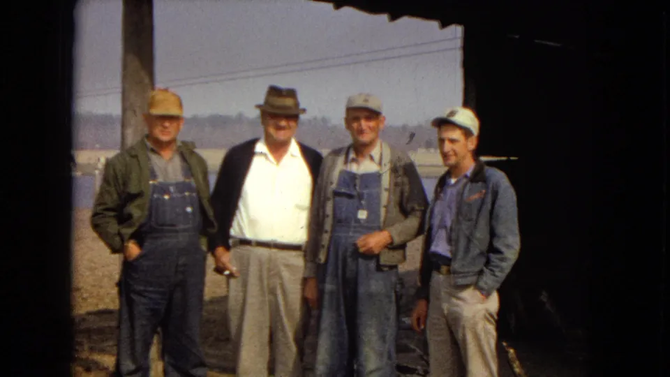 The group remains posed under the shaded roof edge, with three men in denim overalls or work jackets and the central man in a white shirt, dark jacket, and fedora.