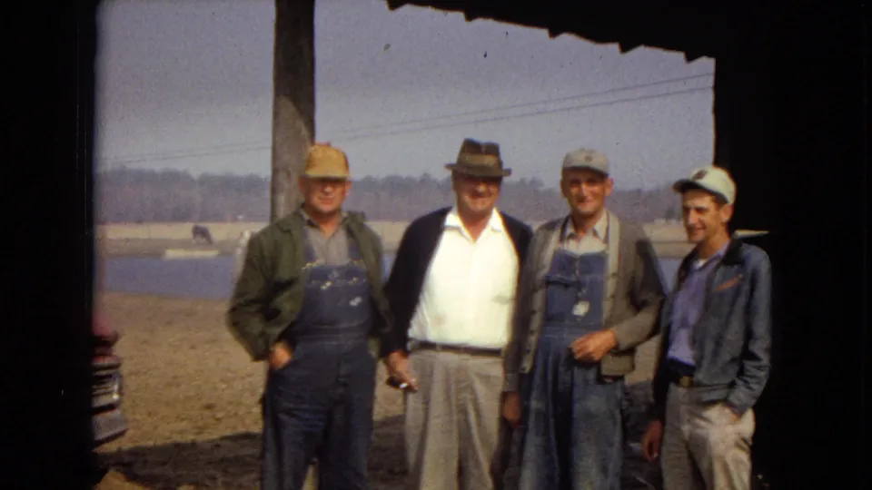Four older men face the camera under the porch roof, with a partial vehicle edge at the left, a body of water in the background, and open dirt ground around them.