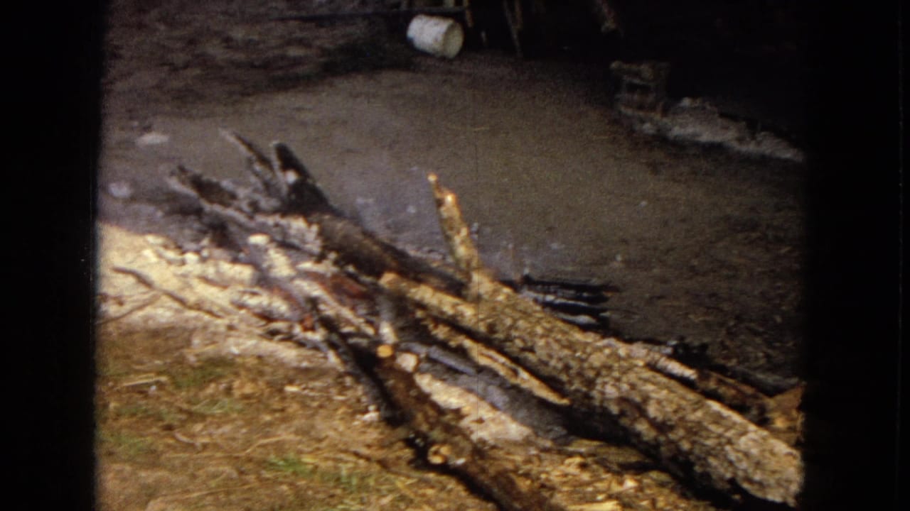1967 archival 8mm frame from the United States showing a close view of charred firewood and glowing embers on dirt ground at night.