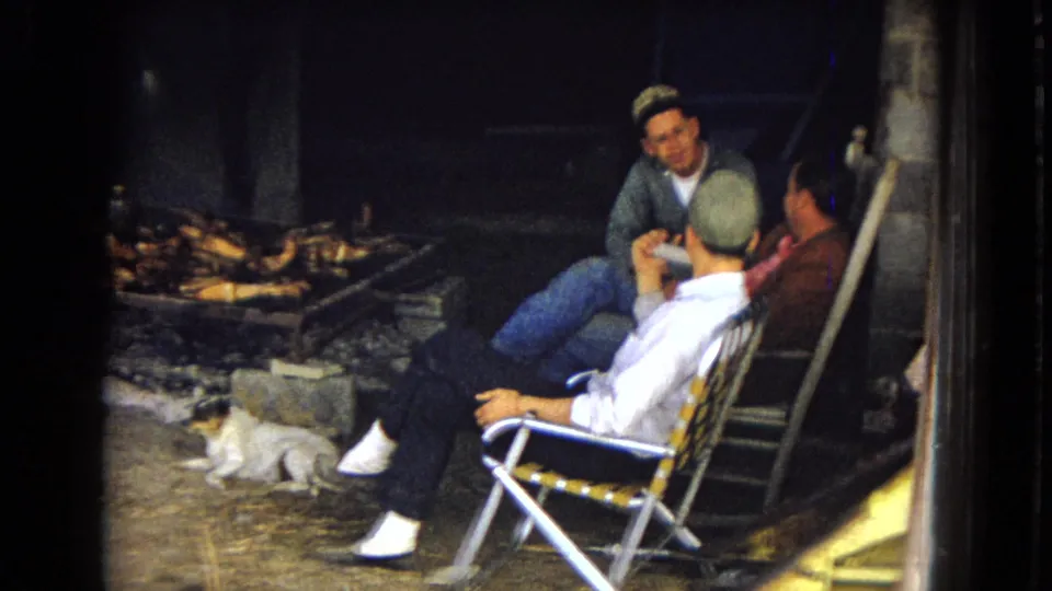 1967 archival 8mm frame from the United States showing three men seated in folding chairs near a nighttime outdoor cook fire, with a small dog resting nearby.