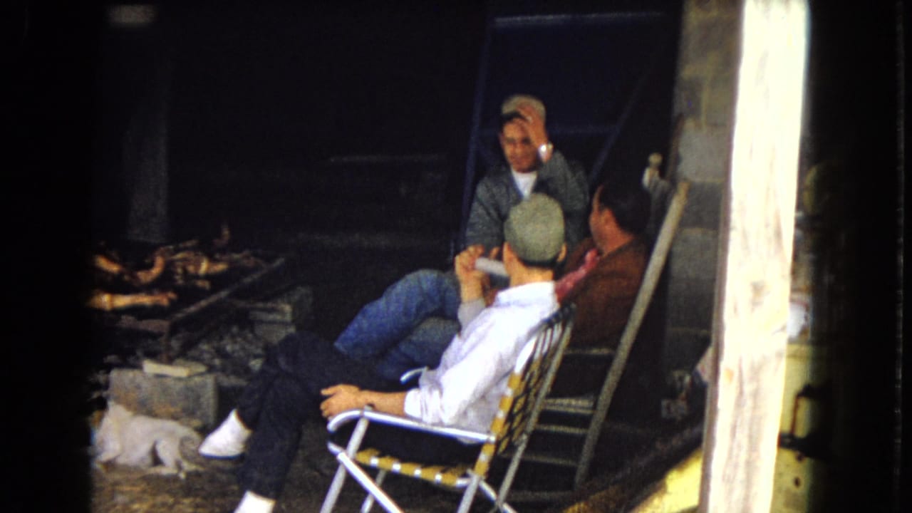 In supported 1967 United States home-movie footage, three men sit casually in folding lawn chairs near a grill area, with one man turned away in a cap and another gesturing while talking.