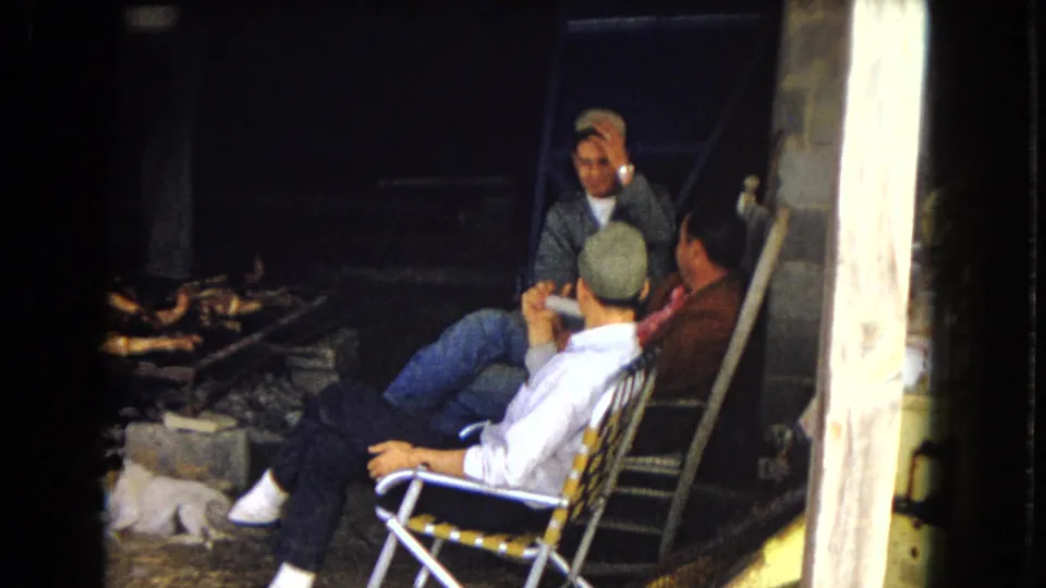 In supported 1967 United States home-movie footage, three men sit casually in folding lawn chairs near a grill area, with one man turned away in a cap and another gesturing while talking.