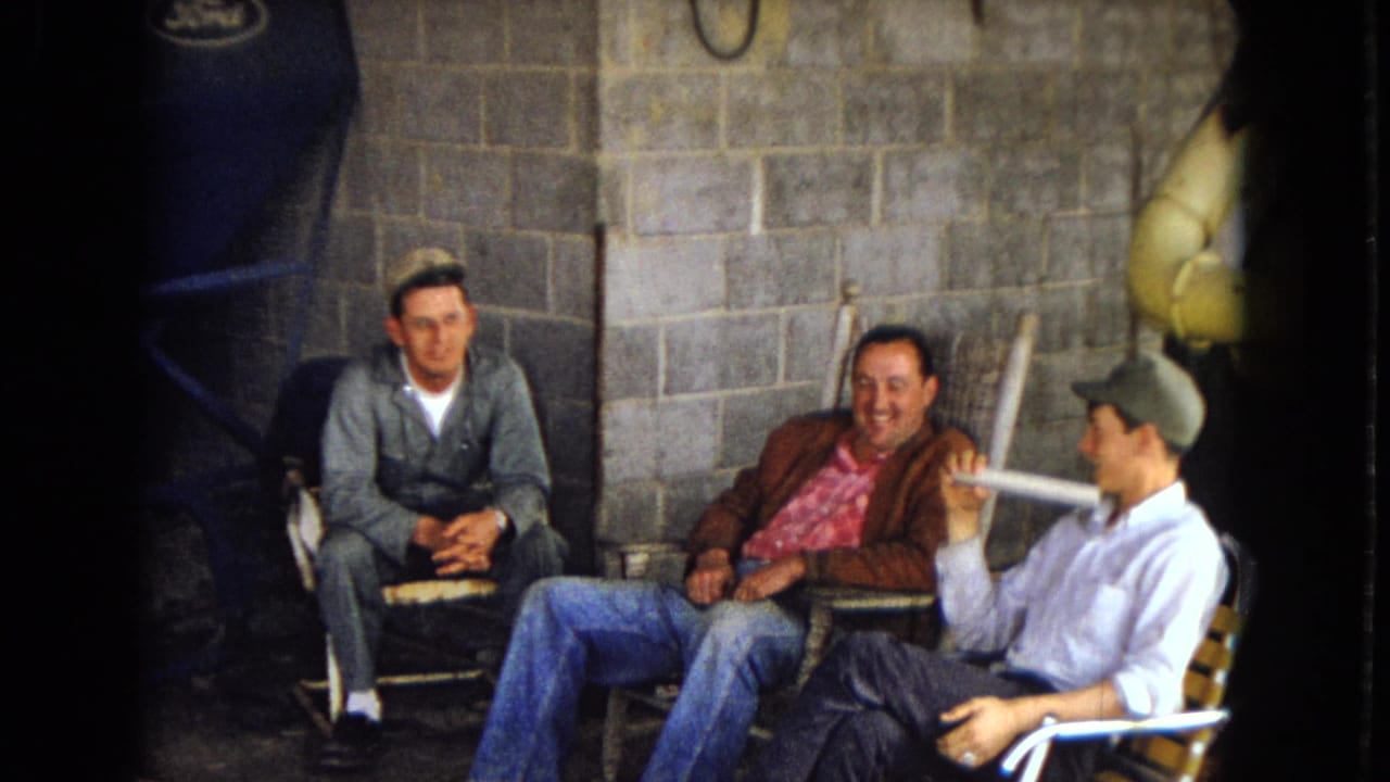 Three men sit against a cinder block wall, smiling and talking in lawn chairs.