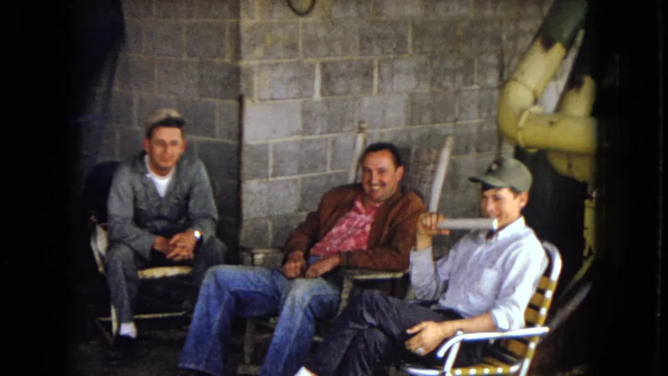 In a casual outdoor or semi-outdoor setting, three men sit close together in folding chairs, smiling beside a cinder block wall with visible pipes at the right edge.