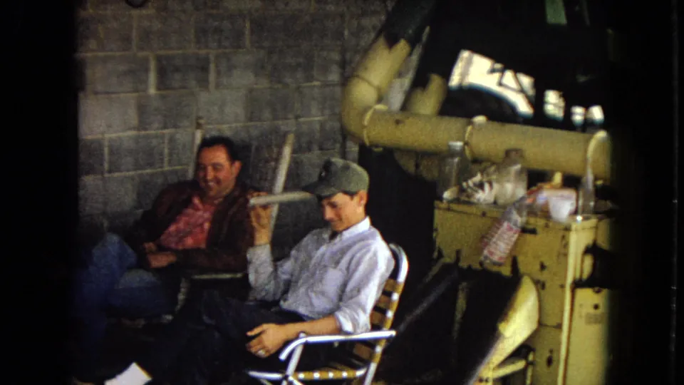 Two seated men are most visible near exposed pipes and barbecue or utility equipment, with one man in a cap holding a rolled paper while another smiles from a chair.
