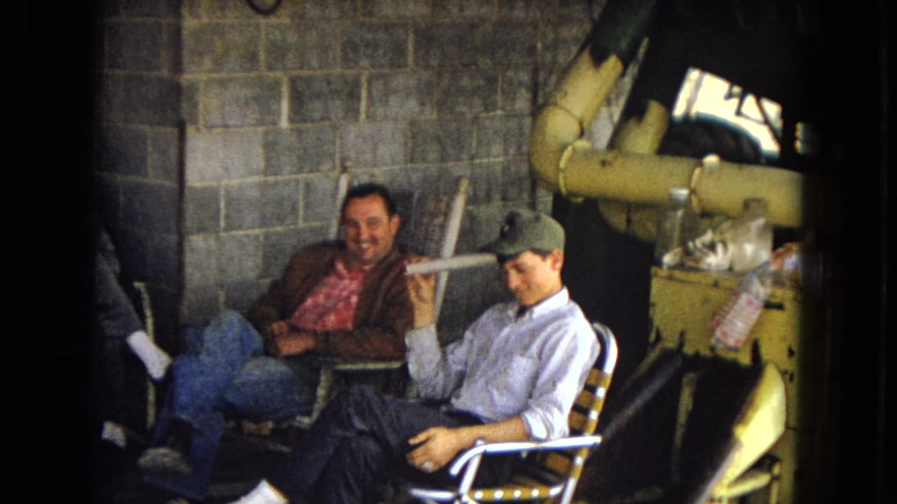 In supplied 1967 archival home-movie footage from Arizona, two adult men sit in folding chairs near cinder block walls and yellow piping while a partially visible third man sits at left.
