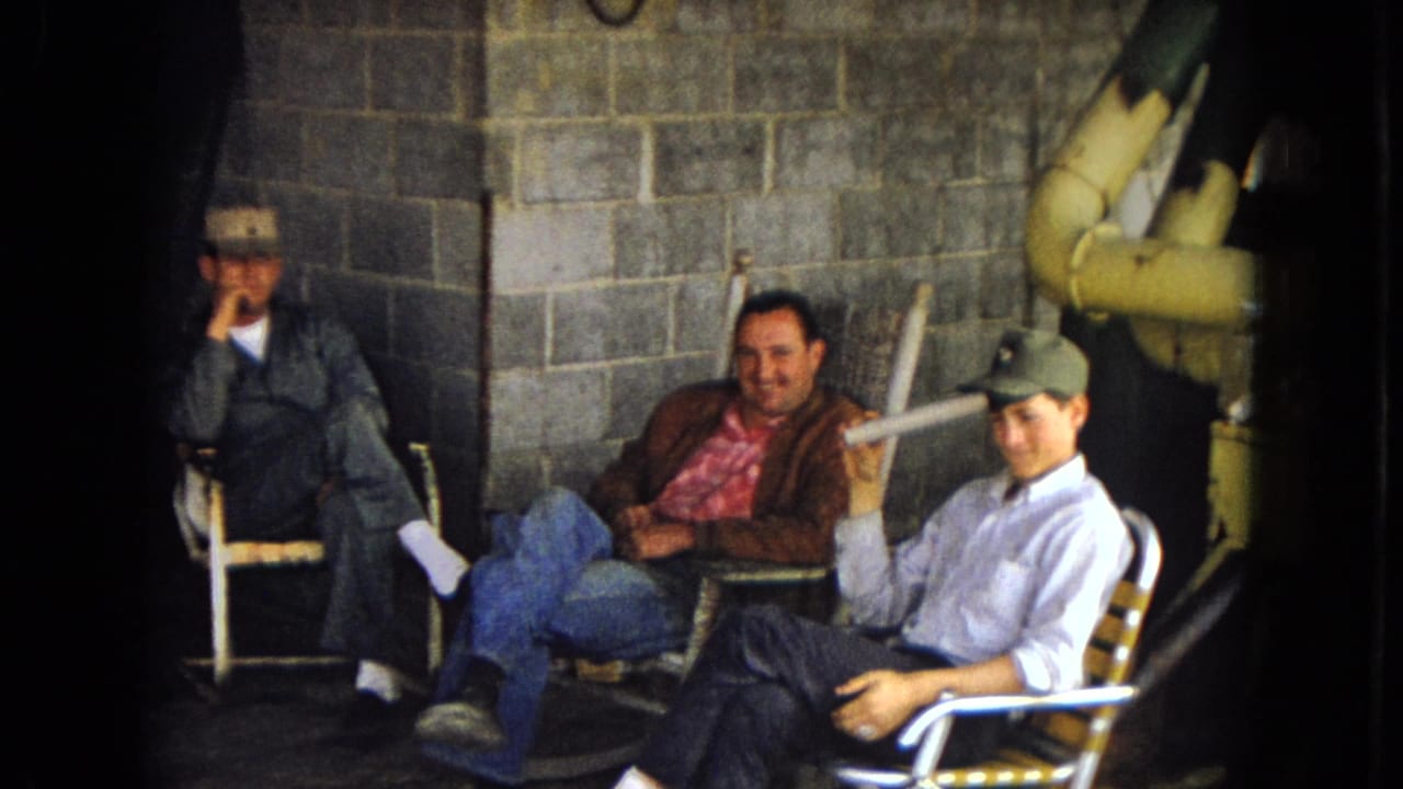Three adult men sit in folding chairs against a cinder block wall, with one man smoking and another smiling.