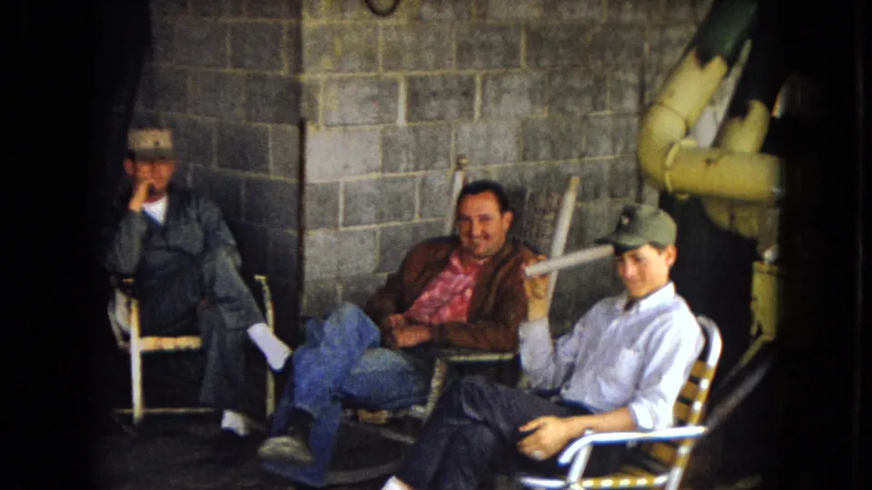 Three adult men appear seated in a dim Arizona utility or work-site room in 1967, including one in a cap holding a cigarette and another smiling in a brown jacket.