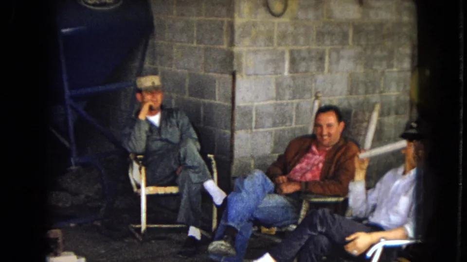 The 1967 home-movie frame shows three adult men seated informally in a dim cinder block utility space, with one man partly cropped at right and industrial-looking equipment around them.