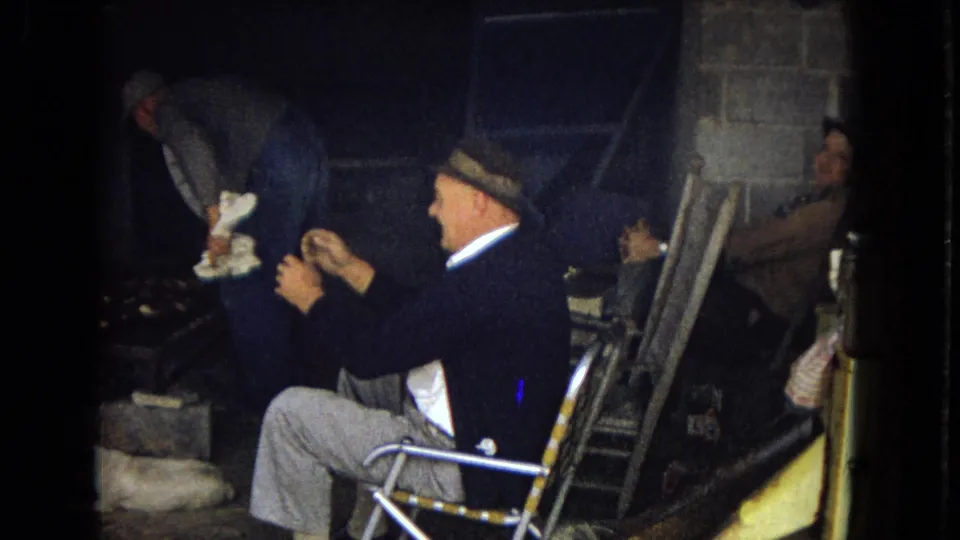 A man in a brimmed hat sits in profile in a folding chair and holds a cigarette inside a dark utility or basement-like room, with another figure partly visible in the background.