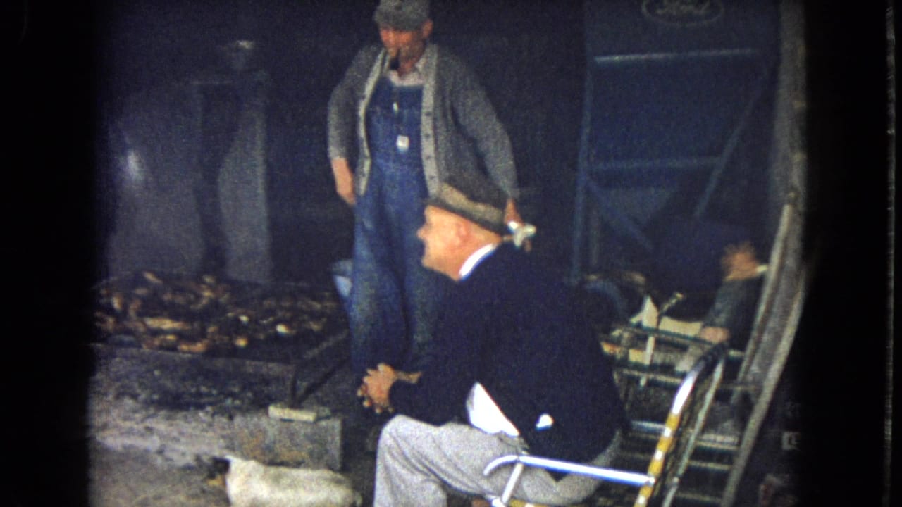 1967 8mm frame showing a man in blue overalls holding or smoking a pipe while a seated older man in a hat faces him in a low-light rustic outdoor area.