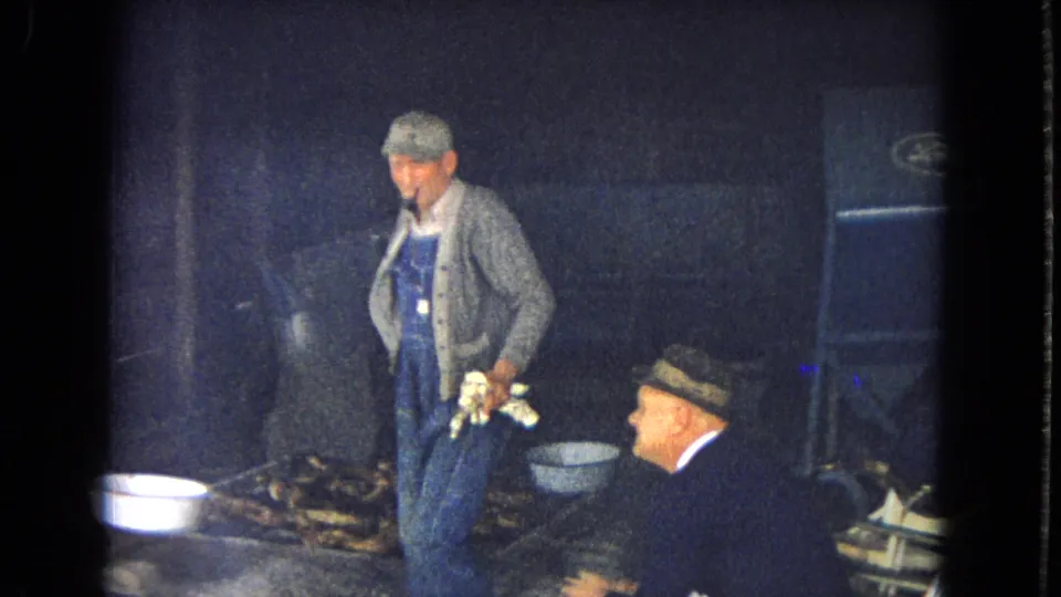 Low-light 1967 home-movie frame showing an older man in overalls and cardigan standing near stacked wood, a white basin, and another older man seated at the edge of the frame.