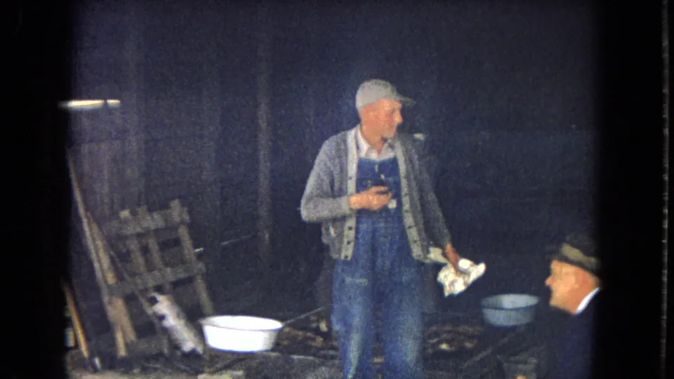 1967 8mm frame from a rustic outdoor scene showing a man in blue overalls holding a pipe and cloth while another older man sits nearby, with basins, stacked wood, and a white dog visible.