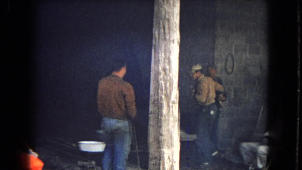 In 1967 Arizona home-movie footage, several adult men in work clothes stand inside or beside a dark industrial setting with cinder block walls, posts, metal basins, and an orange car hood partly visible.