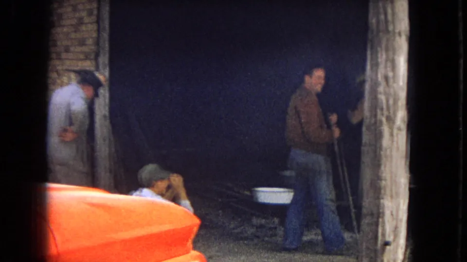 A 1967 Arizona frame shows adult men in hats and jackets gathered near a dark wall with folding chairs, a metal basin, long poles or sticks, and the orange hood of a car in the foreground.