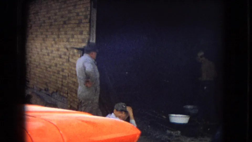 This 1967 Arizona home-movie frame shows men near a brick wall and shadowed industrial space, with one man standing, another seated low, and an orange car hood dominating the foreground.