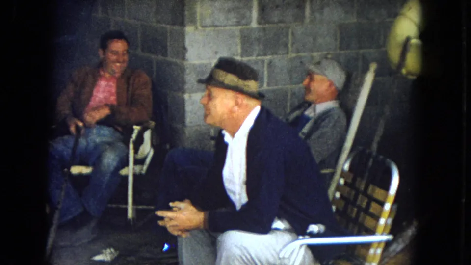 In a clearer 1967 Arizona frame, three adult men sit on folding chairs near a cinder block wall, wearing hats, jackets, jeans, and a cardigan while talking during a break.