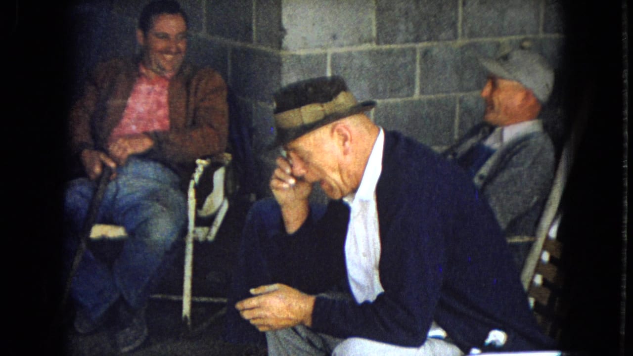 In 1967 home-movie footage identified as Arizona, older men sit against a block wall while a man in a fedora and dark cardigan gestures during conversation.