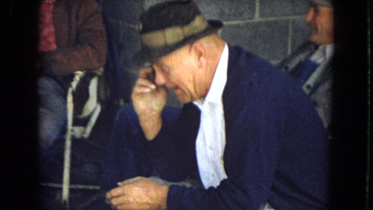 A man in a fedora leans forward with his hand near his face while another man sits behind him.