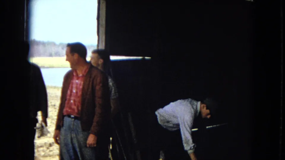 Inside a shadowed barn or shed, a man in a brown jacket stands near daylight at the doorway while another man bends toward a low cooking area.