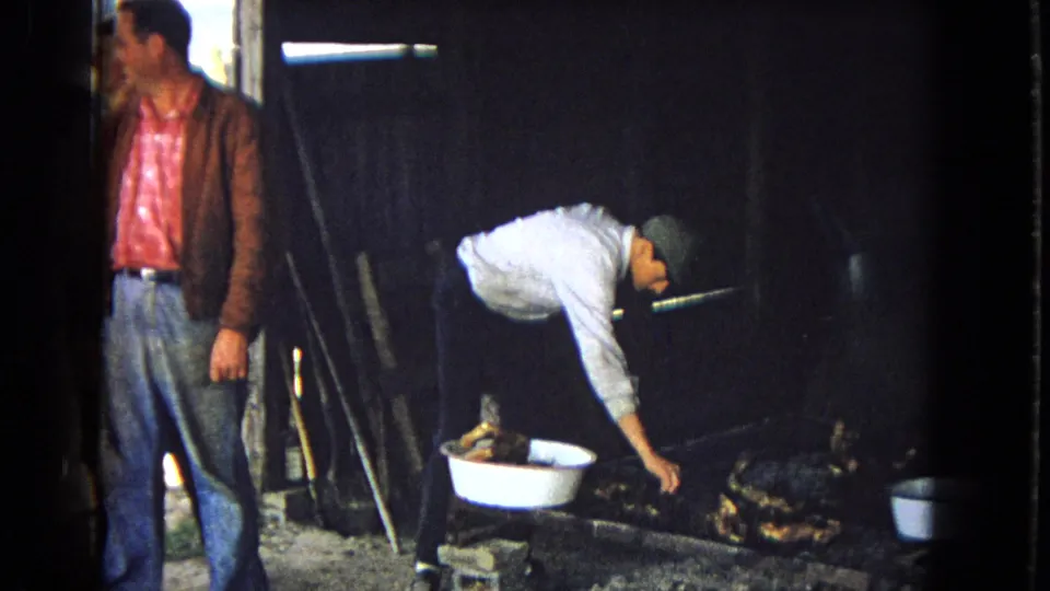 A man bends toward a small fire or cooking pit beside a white basin while another man in a brown jacket stands in the dim barn-like interior.