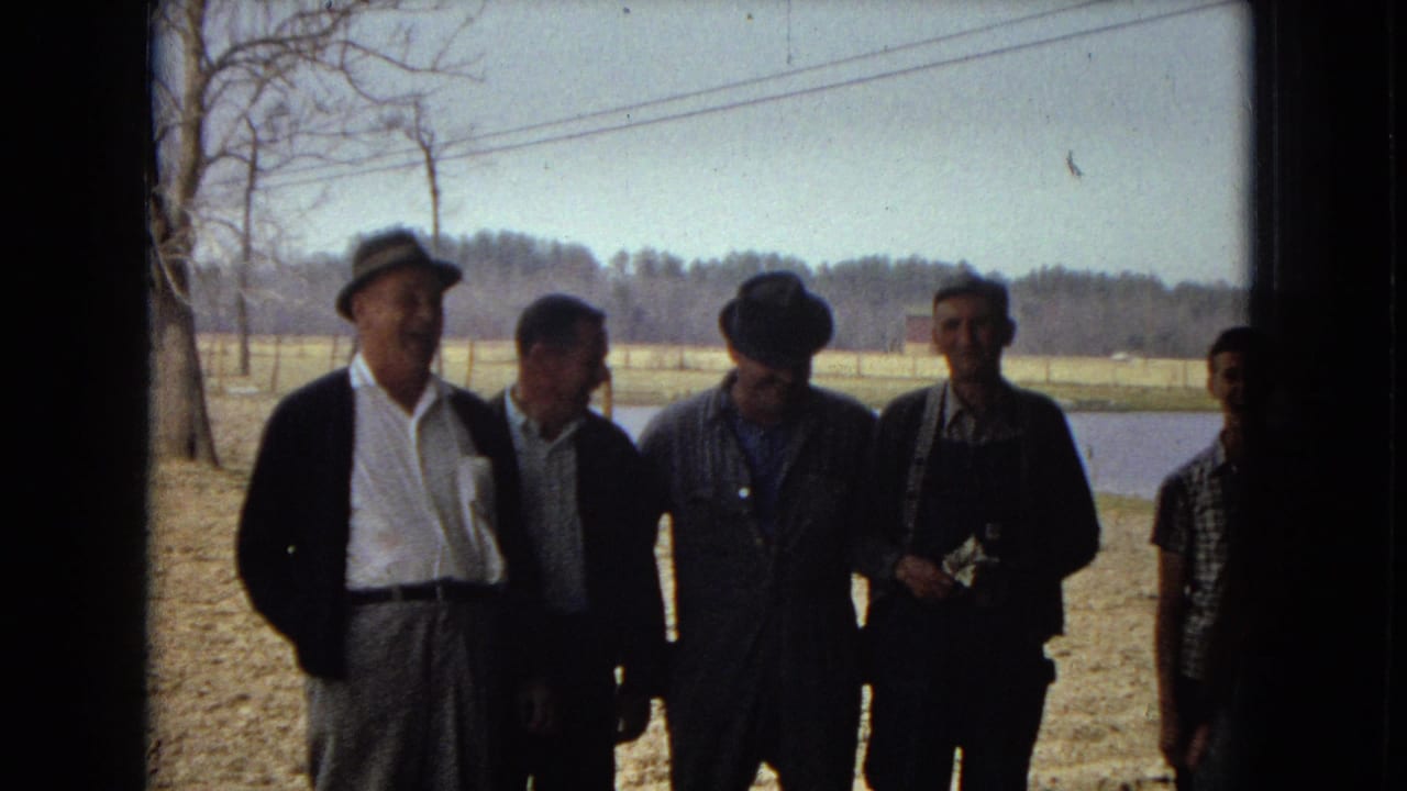 A group of adult men stand outdoors in casual 1960s clothing near bare trees, open ground, and a pond or small body of water in metadata-supported United States home-movie footage from 1967.