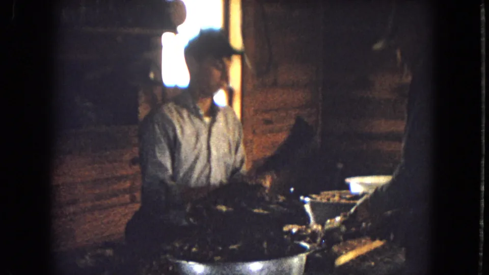 A man in a cap sits beside a table inside the dim cabin as another adult stands nearby handling food or cookware, with log walls and a bright window visible behind them.
