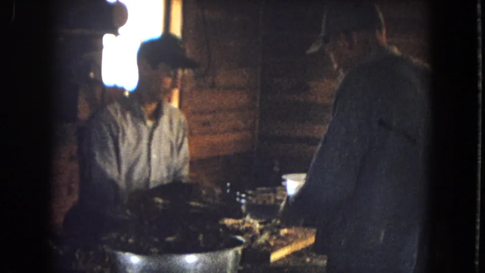 Two adult men remain around the cabin table, with one seated near a large bowl and another standing in partial silhouette while food or utensils are handled in low indoor light.