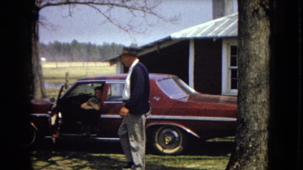 In the supplied 1967 United States home-movie footage, a man in a fedora and dark jacket walks past a maroon sedan while another adult sits inside the open car door near farm buildings and trees.