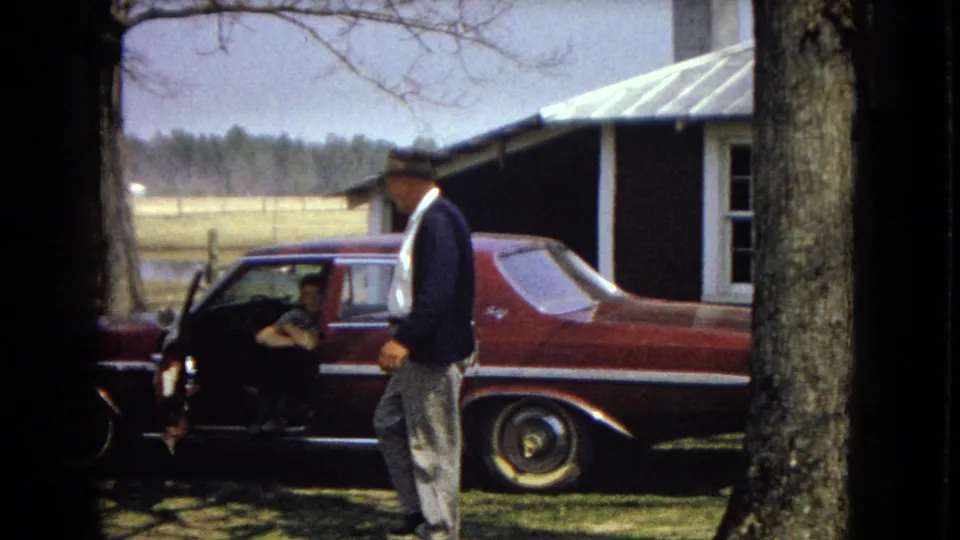 In the supplied 1967 United States home-movie footage, a man in a fedora and dark jacket walks past a maroon sedan while another adult sits inside the open car door near farm buildings and trees.